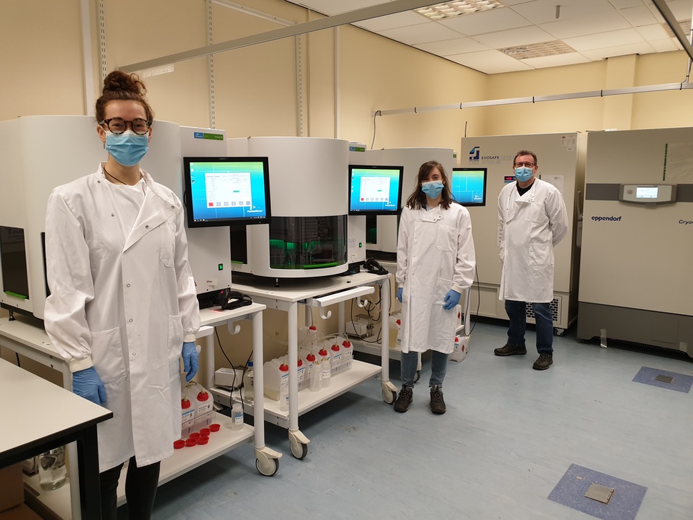 Photograph of a laboratory with three scientists in white coats, a number of DNA sequencing machines and two ultra-low temperature freezers